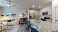 This single-floor residence is designed with a contemporary open-concept layout that seamlessly connects the kitchen, dining, and living areas. The kitchen is outfitted with white shaker-style cabinetry, granite countertops, a subway tile backsplash, and a full suite of stainless steel appliances, including a French door refrigerator, electric range, and over-the-range microwave. A central island with a built-in sink and bar seating serves as both a functional workspace and a casual gathering point. Wood-look flooring extends throughout the main living area, grounding the bright, neutral color palette.

The living area accommodates a comfortable seating arrangement and receives natural light through a large window fitted with blinds and curtains. The bedroom continues the same wood-look flooring, features a ceiling fan, and includes a window that brings in natural light. An expansive walk-in closet off the bedroom provides multi-level wire shelving and ample hanging space. The bathroom presents a wide granite-topped vanity with gray shaker cabinetry, a large mirror, and recessed lighting, maintaining the cohesive, modern aesthetic found throughout the residence.