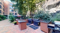 Located at 100 Paterson Plank Rd in Jersey City, NJ, this thoughtfully designed outdoor courtyard offers a welcoming urban retreat set against the backdrop of a classic multi-story red brick building. The ground-level space features a distinctive herringbone-patterned brick pathway that weaves throughout, connecting the courtyard's various functional zones.

Abundant greenery is a defining feature, with mature trees, dense shrubs, flowering plants, and manicured landscaping creating a lush, park-like atmosphere. The courtyard includes multiple seating areas outfitted with round picnic-style tables topped with large umbrellas in both navy and red, providing comfortable shaded spaces for dining or socializing. Wooden benches are also tucked among the plantings for quieter moments of relaxation.

A dedicated outdoor kitchen area features several built-in BBQ grilling stations with dark countertops, ideal for community gatherings and outdoor cooking. Adjacent to this, a section of artificial turf sits just outside large glass-paneled doors leading to an interior fitness facility, blending indoor and outdoor activity seamlessly.

A set of stone stairs with classic black iron railings connects different levels of the courtyard, adding architectural character. The natural rock face visible along one boundary further enhances the unique, nature-integrated character of this well-appointed outdoor space.