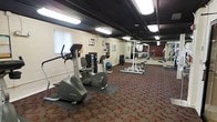 This fitness center offers a practical and functional workout environment designed to accommodate a variety of exercise routines. The space is laid out on a single floor and features durable rubber tile flooring, providing both safety and comfort during workouts. The ceiling is open and industrial in style, with exposed beams and overhead fluorescent lighting that keeps the entire area well-lit. The walls are finished in light-colored concrete block, giving the space a clean and utilitarian aesthetic. Cardio equipment, including a Life Fitness elliptical trainer and a stationary bike, is positioned near the entrance, making them easily accessible. Further into the space, a dedicated strength training area features a weight rack stocked with a full range of plate weights, a barbell station, and a workout bench, ideal for resistance training. A stability ball and additional accessories are also available for functional fitness exercises. Wall-mounted mirrors in the strength area allow users to monitor their form during lifts. A window unit provides climate control, ensuring comfort during workouts year-round. Overall, this fitness center delivers a comprehensive and no-frills workout environment suitable for both cardio and strength training needs.