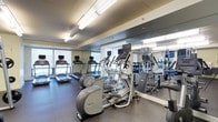 This fitness center is organized into distinct zones designed to accommodate a wide range of training needs. The cardio area is lined with a row of treadmills and elliptical machines, each equipped with individual monitors for entertainment during workouts. Large windows along one wall allow natural light to filter in, creating an open and energizing atmosphere. Floor-to-ceiling mirrors amplify the sense of space and allow users to monitor their form throughout their sessions. The flooring throughout the cardio and strength zones features durable rubber matting, providing safety and comfort underfoot.

The strength training area is well-appointed with a cable machine, a squat rack with an adjustable bench, free weights, and barbells, offering comprehensive options for resistance training. A wall-mounted flat-screen television and mirrored walls keep the space visually engaging.

A separate functional training studio features polished hardwood flooring and a clean, open layout ideal for yoga, stretching, or group fitness. This room is equipped with kettlebells, medicine balls, stability balls, a BOSU trainer, and exercise mats. Large windows provide views to the outdoors, enhancing the bright, airy ambiance. Recessed lighting and a wall-mounted television complete the studio's modern, well-designed interior.