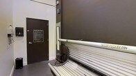 This single-floor tanning facility is purpose-built for personal care and wellness services. The space centers around a professional-grade tanning bed, a full-length unit equipped with multiple UV lamps designed to deliver an even, consistent tan. The room features a dark, moody color palette with deep charcoal walls that contrast against the white trim and lighter flooring, creating a private and focused atmosphere. A small stool is positioned conveniently beside the tanning bed, offering a place to sit while preparing for a session. A posted "Tanning Bed Rules" sign is affixed to the door, ensuring users are informed of proper safety guidelines and usage protocols before entering. The entry door provides a secure, private enclosure for guests during their sessions. The overall layout is streamlined and utilitarian, making efficient use of the available space while keeping the user experience straightforward. The facility appears well-suited for use within a gym, salon, or wellness center, offering clients a convenient and dedicated space for UV tanning services.