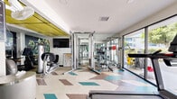 Located in Porter Ranch, Los Angeles, this fitness center offers a thoughtfully designed exercise space across a single floor. The room features a striking chevron-patterned floor in neutral tones accented by bold teal and brown geometric inlays, lending the space a modern, energetic character. A distinctive yellow ceiling panel adds a pop of color overhead, complementing the otherwise clean white walls and creating a visually dynamic environment.

The facility is well-equipped for a comprehensive workout. Cardio equipment includes treadmills, elliptical trainers, and stationary bikes, with a wall-mounted television providing entertainment during sessions. Large floor-to-ceiling mirrors line portions of the walls, enhancing the sense of openness and allowing users to monitor their form. A Hoist cable machine and additional strength training equipment round out the resistance training offerings, while a dedicated rack holds a full set of dumbbells and a separate station stores kettlebells and medicine balls for functional training.

Natural light pours in through large sliding glass doors at one end of the room, which open to an outdoor courtyard with lush greenery and tree views, creating an airy, connected atmosphere. The layout is open and organized, allowing for easy movement between equipment zones throughout the space.