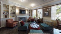 Located at 46 River St in Boston, MA, this single-floor community lounge space offers a warm and inviting atmosphere designed for relaxation and social interaction. The interior is characterized by classic architectural detailing, including crown molding, wainscoting panels along the lower walls, and a tray ceiling that adds a sense of structure and elegance to the room.

At the heart of the lounge sits a white-mantled fireplace flanked by built-in bookshelves filled with books and decorative objects, with a wall-mounted flat-screen television above — creating a comfortable focal point for gatherings. Multiple seating arrangements are thoughtfully distributed throughout, featuring a mix of striped sofas, upholstered armchairs in varied patterns, and teal ottomans surrounding a round central coffee table, encouraging both conversation and leisure.

Large windows dressed with sheer curtains allow generous natural light to fill the space while offering views of the surrounding greenery. An additional seating nook near the windows provides a quiet spot for reading or small group interactions. A built-in beverage station with dark wood cabinetry is conveniently integrated into the space, along with a service window connecting to an adjacent area. The patterned carpeting and warm, earth-toned walls complete the cozy, residential ambiance of this well-appointed community lounge.