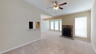 This single-story home offers a thoughtfully designed layout that balances comfort and functionality. The entryway greets residents with rich, dark hardwood flooring, crisp white trim, and neutral textured walls, setting a clean and inviting tone. A built-in HVAC vent and light switch are conveniently positioned near the front door, while an adjacent hallway leads further into the home.

The living area is a standout space, distinguished by soaring vaulted ceilings that enhance the sense of openness and volume. A ceiling fan with a light fixture is centrally mounted, providing both airflow and ambient lighting. Plush, neutral-toned carpeting runs throughout, adding warmth underfoot. A stone-surround fireplace anchors one wall, serving as a natural focal point and lending a cozy, classic character to the room. Large windows along the far wall invite natural light, brightening the space throughout the day.

A pass-through window with a tiled ledge connects the living area to the kitchen, fostering an open, communicative flow between spaces. A pendant light fixture hangs above, highlighting this architectural detail. The overall ambiance is bright, airy, and move-in ready.