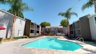 This residential complex is organized around a central outdoor courtyard that serves as the focal point of the community. The courtyard features a generously sized swimming pool with clear blue water, surrounded by an expansive concrete sun deck well-equipped with lounge chairs, shaded umbrella tables, and a barbecue area for outdoor gatherings. Tall palm trees are strategically placed throughout the courtyard, adding a distinctly warm, resort-like atmosphere under the consistently clear, sunny sky.

The multi-building layout consists of two-story structures finished in neutral tones of gray and taupe, with blue accent panels adding a contemporary touch. Private balconies on the upper levels overlook the shared courtyard, providing residents with direct views of the pool and landscaping below. Ground-floor units have walkout access to the patio areas. A pergola structure on one side of the deck provides additional shade, complementing the red market umbrellas positioned around seating clusters. Secured gated entryways with ornamental metal fencing define pathways between buildings, with container plantings and maintained shrubs enhancing the landscaped grounds. The overall ambiance is open, sun-filled, and oriented toward communal outdoor living.