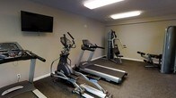 This fitness room is a well-appointed exercise space designed to support a range of workout routines. The room features rubber flooring suited for gym use, providing durability and comfort underfoot. Overhead fluorescent lighting ensures the space is brightly and evenly lit throughout. The neutral-toned walls contribute to a clean, focused atmosphere.

The room is equipped with multiple cardio machines, including two treadmills and an elliptical trainer, allowing for varied cardiovascular workouts. A multi-station strength training unit is positioned toward the back of the room, offering options for resistance and weight-based exercises. A wall-mounted flat-screen television is installed near the entrance, providing entertainment during workouts.

The layout is functional and efficiently organized, with equipment arranged to allow adequate movement between machines. The space is designed for practical, everyday fitness use, making it a convenient option for those seeking an accessible workout facility.