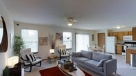 This two-floor home offers a clean, well-organized layout designed for comfortable everyday living. The main living area is bright and airy, featuring large windows with horizontal blinds and vertical slat coverings that allow abundant natural light to fill the space. The living room is thoughtfully arranged with a plush gray sofa, accent chairs with bold black-and-white cushions, and an oval coffee table as the central focal point. Decorative elements, including wall art and a tall dried floral arrangement, add character and warmth to the neutral color palette of white walls and light carpeted flooring. A ceiling fan with a light fixture provides both comfort and illumination overhead. Adjacent to the living area, a dining space is visible, featuring a light-toned table and chairs, seamlessly connected to the living room to create an open, sociable floor plan. A door near the dining area leads to an additional room or exterior space, adding to the functional flow of the layout. The overall ambiance is calm and inviting, with a consistent neutral aesthetic that provides a versatile backdrop suitable for a variety of interior styles. The two-floor configuration further expands the living space, offering separation between communal and private areas of the home.