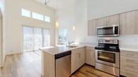 This single-floor unit showcases a thoughtfully designed open-concept layout anchored by a striking kitchen with dramatically tall ceilings. The kitchen is equipped with stainless steel appliances, including a refrigerator, electric range, built-in microwave, and dishwasher. Light wood-toned cabinetry extends nearly to the ceiling, maximizing storage, while crisp white countertops and a peninsula with a built-in sink provide generous workspace and informal dining options. Pendant lighting hangs above the peninsula, complementing the ceiling fan mounted high above the living area. Warm hardwood-style flooring runs throughout the main living spaces, lending a cohesive and inviting feel.

Large windows and sliding glass doors with vertical blinds flood the open living area with natural light and offer views to the outdoors. The bedroom is a calm, neutral space featuring soft carpet flooring and two windows that bring in additional daylight. The bathroom maintains the unit's consistent aesthetic with wood-toned vanity cabinetry, a light countertop with an undermount sink, rich hardwood-style flooring, and a full tub-shower combination. Clean white walls throughout reinforce the modern, minimalist character of the space.