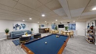 This well-appointed single-floor amenity space is designed to accommodate a variety of social and recreational activities, combining comfort and functionality in a cohesive, modern aesthetic. The layout is anchored by a full-sized billiards table with a blue felt surface, positioned at the heart of the room and surrounded by ample open floor space for gameplay and circulation.

A comfortable lounge area features a blue-gray sofa, sleek accent chairs with gold-toned frames, a marble-top coffee table, and a boldly colored abstract canvas that adds a vibrant focal point to the wall. Lush potted greenery is placed throughout, contributing a fresh, lively atmosphere.

Toward the rear of the space, a dedicated dining area includes multiple round tables with rattan-style chairs, ideal for casual gatherings. A flat-screen television is mounted on the wall nearby, and natural light filters in through several windows overlooking surrounding greenery.

A stylish bar counter with a stone-topped surface and dark fluted panel detailing serves as a social hub, accompanied by bar-height seating. A rectangular mirror above the counter enhances the sense of depth in the space.

A quieter alcove accessed through French doors provides a private work or study corner, complete with a desk and chair. Throughout, recessed lighting and a drop ceiling maintain a clean, polished finish, while gray wood-look flooring ties the entire space together.
