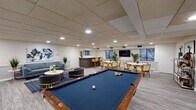 This well-appointed single-floor amenity space is designed to accommodate a variety of social and recreational activities, combining comfort and functionality in a cohesive, modern aesthetic. The layout is anchored by a full-sized billiards table with a blue felt surface, positioned at the heart of the room and surrounded by ample open floor space for gameplay and circulation.

A comfortable lounge area features a blue-gray sofa, sleek accent chairs with gold-toned frames, a marble-top coffee table, and a boldly colored abstract canvas that adds a vibrant focal point to the wall. Lush potted greenery is placed throughout, contributing a fresh, lively atmosphere.

Toward the rear of the space, a dedicated dining area includes multiple round tables with rattan-style chairs, ideal for casual gatherings. A flat-screen television is mounted on the wall nearby, and natural light filters in through several windows overlooking surrounding greenery.

A stylish bar counter with a stone-topped surface and dark fluted panel detailing serves as a social hub, accompanied by bar-height seating. A rectangular mirror above the counter enhances the sense of depth in the space.

A quieter alcove accessed through French doors provides a private work or study corner, complete with a desk and chair. Throughout, recessed lighting and a drop ceiling maintain a clean, polished finish, while gray wood-look flooring ties the entire space together.