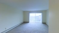This single-floor residential space offers a clean, functional layout centered around a generously sized living area. The room is finished with neutral, light-toned walls and wall-to-wall carpeting in a soft, muted hue, contributing to a calm and understated atmosphere throughout.

A prominent sliding glass door serves as the focal point of the main living area, framed by vertical blinds and flanked by large windows that allow an abundance of natural light to fill the room. The glass door opens to a balcony, providing outdoor access and views of the surrounding landscape, which includes open green space and adjacent multi-story buildings visible in the background.

Baseboard heating units run along the perimeter of the room, offering an efficient heating solution integrated discreetly into the space. The smooth, white ceilings are standard in height and contribute to the room's bright, open feel.

The overall design is straightforward and practical, with a neutral color palette that lends itself well to a variety of furnishing styles. The abundance of natural light, outdoor access via the balcony, and open floor plan make this a comfortable and versatile single-floor living space.