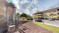 Located at 980 Courthouse Rd, this single-floor community space is part of a well-established residential complex characterized by its warm yellow exterior buildings and open, welcoming grounds. The outdoor area serves as a central gathering point, featuring a dedicated playground complete with climbing structures, slides, and play equipment set on a wood chip surface — ideal for recreational use. Mature trees and lush greenery are interspersed throughout the property, providing natural shade and a pleasant, park-like atmosphere. The grounds are neatly maintained, with manicured lawns and paved pathways connecting the various areas of the complex. A chain-link fence borders portions of the outdoor space, delineating the playground from the adjacent parking area. The multi-story residential buildings in the background showcase covered balconies and large windows, contributing to an airy, open feel across the community. The property enjoys generous natural light and open sky views, further enhancing the sense of space. Overall, 980 Courthouse Rd presents a community-oriented environment with thoughtfully designed outdoor amenities that foster an active and connected lifestyle.