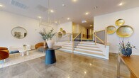 This elegantly designed building lobby makes a striking first impression with its expansive polished stone tile flooring that reflects the abundant natural light streaming in through floor-to-ceiling windows along the street-facing facade. The open, airy layout is anchored by a central seating area furnished with warm amber velvet chairs, a sculptural round coffee table, and a woven area rug, creating an inviting gathering space. A bold geometric brass chandelier overhead adds a refined, modern touch to the bright white ceiling fitted with recessed lighting throughout.

A prominent central staircase serves as an architectural focal point, featuring light wood treads, crisp white risers, and sleek black iron balusters capped with natural wood handrails — a design detail that balances contemporary style with warmth. Decorative circular wall niches accented in warm gold tones line the staircase wall, adding visual depth and character.

A dedicated workspace area near the staircase includes a mid-century modern wood desk, while additional seating near the window wall provides a comfortable lounge zone. Curated wall art and sculptural décor pieces complete the sophisticated aesthetic. The overall ambiance is modern, welcoming, and professionally appointed, reflecting thoughtful attention to both function and design.