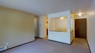 This single-floor residence offers a thoughtfully arranged open layout designed for comfortable everyday living. The main living area is finished with plush wall-to-wall carpeting in a neutral tone, creating a warm and inviting atmosphere. A distinctive architectural feature of the space is the built-in pass-through window set within a white structural partition, which connects the living area to the kitchen — a practical design element that promotes an open feel while maintaining a defined separation between spaces. The kitchen, visible through the pass-through, features dark cabinetry and a ceiling light fixture, giving it a cozy and functional character. The partition itself adds visual interest to the layout, breaking up the open floor plan while allowing light to flow between areas. A hallway accessible from the living space leads further into the residence, hinting at additional rooms beyond. The walls and ceilings are finished in crisp white and soft neutral tones, contributing to a clean, understated aesthetic throughout. Dark baseboards provide a subtle contrast against the light carpeting and walls, adding a grounding detail to the overall design. The space is well-lit and presents a move-in ready environment with a straightforward, functional floor plan.