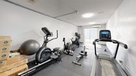 Located in Miami, FL, this dedicated exercise room offers a focused fitness environment within a compact, single-room layout. The space features white walls and a textured ceiling, lending it a clean, functional aesthetic. Dark interlocking foam flooring tiles cover the entire floor area, providing cushioning and stability suitable for a variety of workout activities.

The room is equipped with a well-rounded selection of cardio and strength machines, including a treadmill, a rowing machine, an elliptical trainer, a spin bike, and a stair climber. Free weights and a stability ball round out the equipment offerings. A designated stretching and yoga area is defined by exercise mats laid out near the sliding glass door, creating a quieter zone for low-impact workouts.

Climate control is handled by a wall-mounted ductless split air conditioning unit, ensuring a comfortable workout environment year-round — an essential feature in the Miami heat. Natural light enters through a sliding glass door at one end of the room, which also provides direct outdoor access. Overhead flush-mount light fixtures provide consistent artificial lighting throughout the space. A separate entry door is located at the opposite end of the room, offering additional access. The overall layout is straightforward and efficient, maximizing the usable floor space for fitness activities.
