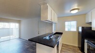 Located at 1845 N Broadway in Escondido, this single-story unit offers a functional and well-appointed layout. The kitchen serves as a central feature of the space, showcasing dark speckled granite countertops that contrast cleanly with bright white cabinetry above and below. A peninsula-style counter extends into the living area, creating an open, connected feel between the kitchen and the adjacent space. The double-basin sink is positioned along the peninsula, adding practicality to the layout. Upper cabinets provide ample storage, while a textured ceiling adds dimension overhead. Wood-style plank flooring runs throughout, offering a warm and durable surface underfoot. Natural light filters into the space through windows fitted with blinds, contributing to a bright and airy atmosphere. Neutral wall tones complement the overall design, keeping the interior feeling clean and versatile. A doorway visible from the kitchen provides easy access to additional areas of the unit. The open layout and cohesive finishes make this a practical and comfortable single-floor living space.