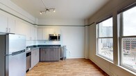 This single-floor space combines classic architectural character with modern updates, creating a comfortable and functional living environment. The open layout seamlessly connects the kitchen and living area, maximizing the sense of space throughout.

The kitchen is well-appointed with contemporary two-tone cabinetry — crisp white upper cabinets paired with warm wood-toned lower cabinets — and dark granite-style countertops. Stainless steel appliances, including an over-the-range microwave and a gas cooktop, add a sleek, modern touch. Track lighting on the ceiling provides focused task illumination across the kitchen workspace.

A defining architectural feature is the decorative green painted border that runs along the upper perimeter of the walls, lending a distinctive vintage charm that nods to the building's heritage. A classic cast-iron radiator stands as a further reminder of the building's history, while contributing to the overall character of the space.

Light hardwood-style flooring runs throughout, adding warmth and continuity. A large, double-hung window floods the space with natural light and frames views of the surrounding urban skyline, reinforcing the city-connected atmosphere of this well-designed interior.