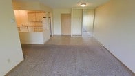 This single-floor unit offers an open, functional layout that combines the living area and kitchen into one cohesive space. The living area is finished with wall-to-wall carpeting, which transitions to tile flooring near the entryway and kitchen zone, providing a clear visual distinction between spaces.

The kitchen is positioned to the left and accessible via a pass-through opening with a raised countertop, creating a semi-open connection to the main living space. Light wood cabinetry lines the upper and lower walls of the kitchen, offering ample storage, while a white refrigerator is visible at the rear. The countertops are clean and functional, suited for everyday use.

The entryway features a wood-toned front door and is flanked by a generously sized closet fitted with mirrored sliding doors, adding both storage and a sense of depth to the space. Overhead, a flush-mount ceiling light provides ambient illumination throughout the main area. The walls are finished in a neutral, light beige tone, contributing to a bright and understated atmosphere throughout the unit.