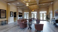 This sophisticated single-floor gathering space exudes a refined, welcoming ambiance anchored by its most striking architectural feature: soaring coffered ceilings with deep, geometric box molding that add drama and grandeur to the interior. Elegant pendant and chandelier-style light fixtures hang at varying heights, casting warm, inviting light throughout the room.

The main lounge area is generously proportioned, furnished with plush upholstered club chairs and sofas in rich earth tones, arranged around a sleek glass-top coffee table to encourage conversation and relaxation. Wide-plank hardwood-style flooring runs throughout, grounding the warm color palette of tans, browns, and deep reds.

Tall white-trimmed windows and sets of French doors line the perimeter, flooding the space with abundant natural light and offering views of the outdoors. White decorative columns frame the open floor plan, adding a classic architectural touch without disrupting the sense of openness.

A secondary seating or dining area is visible deeper in the space, providing additional functionality. A built-in media wall or display feature is visible to one side, enhancing the room's versatility. The overall atmosphere is polished yet comfortable, making this space well-suited for both casual gatherings and more formal social occasions.