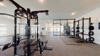 This single-level fitness facility is thoughtfully designed to accommodate a comprehensive range of workout activities across several distinct functional zones. The space features vaulted ceilings with exposed industrial-style ductwork, recessed lighting, and rows of clerestory windows along the upper walls that fill the interior with natural light while maintaining privacy.

The main training floor is finished with durable rubber flooring and houses a robust collection of professional-grade equipment, including a multi-station cable machine by Life Fitness, adjustable benches, a power rack with weight storage, and a full dumbbell rack with accompanying benches. A separate cardio zone accommodates multiple Life Fitness treadmills, elliptical trainers, and a stationary bike, complemented by wall-mounted flat-screen televisions for an engaging workout experience.

A dedicated studio area is enclosed by sleek black-framed glass partition walls, featuring floor-to-ceiling mirrors, rubber mat flooring, and additional strength equipment including a Hammer Strength machine. The white walls and clean architectural lines throughout create a bright, modern aesthetic, giving the facility a polished, professional atmosphere suited for serious athletic training.