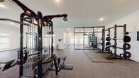This single-level fitness facility is thoughtfully designed to accommodate a comprehensive range of workout activities across several distinct functional zones. The space features vaulted ceilings with exposed industrial-style ductwork, recessed lighting, and rows of clerestory windows along the upper walls that fill the interior with natural light while maintaining privacy.

The main training floor is finished with durable rubber flooring and houses a robust collection of professional-grade equipment, including a multi-station cable machine by Life Fitness, adjustable benches, a power rack with weight storage, and a full dumbbell rack with accompanying benches. A separate cardio zone accommodates multiple Life Fitness treadmills, elliptical trainers, and a stationary bike, complemented by wall-mounted flat-screen televisions for an engaging workout experience.

A dedicated studio area is enclosed by sleek black-framed glass partition walls, featuring floor-to-ceiling mirrors, rubber mat flooring, and additional strength equipment including a Hammer Strength machine. The white walls and clean architectural lines throughout create a bright, modern aesthetic, giving the facility a polished, professional atmosphere suited for serious athletic training.