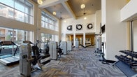 This Wellness Center in Houston, TX (77003) offers a comprehensive, single-floor fitness facility designed to support a wide range of workout routines. The space is architecturally distinguished by soaring double-height ceilings, warm gold accent walls, and an expansive wall of floor-to-ceiling windows that flood the interior with abundant natural light and provide open views of the surrounding courtyard and buildings beyond.

The main fitness floor is generously proportioned, accommodating a robust selection of Technogym strength and resistance machines arranged in an organized layout across patterned, multi-tone carpeting. Cardio equipment, including several treadmills with integrated interactive screens, is positioned directly in front of the large window wall, allowing users to enjoy outdoor views during their workouts.

A dedicated functional training area features medicine balls, stability balls, a cable/squat rack, wall mirrors, and benches, providing space for dynamic and free-form exercise. A dumbbell rack with a full range of free weights is also available, complemented by wall-mounted televisions throughout the space.

The overall ambiance is modern and motivating, with pendant and recessed lighting, contemporary abstract artwork on the walls, and a clean, open layout that ensures ample circulation space between equipment stations. A separate room accessible via glass doors appears to offer additional training or studio space.