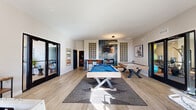 Located at 4501 E Grant Rd in Tucson, Arizona, this single-floor game room is a well-appointed recreational space designed for social entertainment. The room features light hardwood-style flooring throughout, recessed lighting, and high ceilings that lend an open, airy atmosphere. Large windows and glass-paneled doors allow abundant natural light to flood the interior, offering views of the vibrant outdoor surroundings.

The space is centered around a pool table with a bold blue felt surface, complemented by a shuffleboard table and a foosball table. An arcade-style basketball shooting game adds to the variety of entertainment options available. A wall-mounted television is set against a striking teal tile accent wall flanked by decorative breeze-block panels, creating a stylish focal point. Bar-height seating with bistro-style chairs provides a casual gathering spot nearby.

Curated artwork adorns the walls, adding color and personality to the space. Glass double doors connect the game room to adjacent amenity areas, including what appears to be a fitness center and a co-working or lounge space, enhancing the overall connectivity of the facility. The combination of modern design elements and diverse gaming options makes this space a dynamic hub for recreation and community engagement.