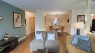 This single-floor space showcases an open-concept design that seamlessly connects the living and dining areas, creating a cohesive and functional layout. The living area features warm hardwood flooring that extends throughout, complemented by neutral beige walls and an accent wall in a muted teal, adding a touch of color and visual interest to the space.

The living area is furnished with modern grey modular seating accented by light blue patterned throw pillows, offering a casual and inviting atmosphere. A hallway visible from the main living area leads to additional rooms, flanked by white-paneled doors and continuing the warm hardwood flooring throughout.

The adjacent dining area is anchored by a round wooden dining table surrounded by matching wooden chairs, beneath a classic pendant light fixture that provides warm, focused illumination. A display cabinet in the background adds both storage and decorative appeal to the dining space.

Natural light flows through the space, highlighting the consistent flooring and clean architectural lines. The overall ambiance is warm, understated, and practical, with a cohesive color palette of neutral tones and natural wood finishes that lend the space a timeless, welcoming character.