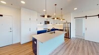 Located at 2913 W Leigh St in Richmond, Virginia, this single-floor unit showcases a thoughtfully designed contemporary interior with high-end finishes and an open, airy layout.

The kitchen serves as a visual centerpiece, featuring bold two-tone cabinetry with cobalt blue lower cabinets contrasted against crisp white upper cabinets that extend to the ceiling for maximum storage. A large island with a light quartz countertop and matte black sink faucet anchors the space, while stainless steel appliances, white subway tile backsplash, and a series of stylish pendant lights complete the modern aesthetic. Light wood-style flooring flows throughout the main living areas.

The living area is defined by expansive floor-to-ceiling grid windows and glass doors that open to a balcony, offering elevated views of the surrounding Richmond neighborhood and flooding the space with natural light.

Bedrooms feature plush carpeting, ceiling fans, and their own floor-to-ceiling window walls with direct balcony access, creating bright, inviting retreats. The bathrooms are finished with distinctive terrazzo flooring, blue-toned vanities with dual sinks, matte black hardware, and white tile surrounds in both the walk-in shower and tub/shower combinations.