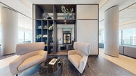 This upscale amenity lounge is a generously proportioned, single-floor space that exemplifies contemporary design at its finest. The layout flows seamlessly from an entry corridor—finished in richly veined dark marble tile and flanked by sculptural décor accents—into a series of open, interconnected seating and social areas.

Throughout the main lounge, expansive floor-to-ceiling glass windows command sweeping panoramic views of the city skyline and waterways below, bathing the interior in abundant natural light. Striking cylindrical columns punctuate the open plan, lending architectural character while subtly defining distinct seating zones. A patterned area rug in muted burgundy and mauve tones anchors the central lounge area, which is furnished with plush sectional sofas and velvet accent chairs in a refined blue-gray palette.

A built-in shelving unit with dark wood framing serves as a design focal point near the entry, displaying curated decorative objects and greenery. Adjacent areas feature light hardwood flooring, a wet bar with stone countertops, and a mounted display screen, catering to both social gatherings and private events. Coffered ceilings with recessed and cove lighting add warmth and depth throughout. The overall ambiance is polished, welcoming, and distinctly elevated—ideal for residents seeking a premium communal retreat.