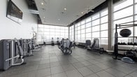 This contemporary fitness center offers a bright and energizing workout environment designed to meet a wide range of exercise needs. The space is defined by an expansive wall of floor-to-ceiling windows that floods the interior with natural light and provides sweeping views of the surrounding cityscape, creating an inspiring atmosphere for any workout routine.

The facility is outfitted with a comprehensive selection of professional-grade equipment, including strength training machines, rowing machines, free weights, and cardio equipment such as ellipticals and upright bikes. The machines are thoughtfully arranged to allow ample space for movement and safe usage throughout the floor.

Underfoot, large-format gray tile flooring provides a durable, easy-to-maintain surface suited for a high-traffic fitness environment. Overhead, a drop ceiling fitted with recessed lighting and ceiling fans ensures consistent illumination and comfortable air circulation throughout the space. The overall design strikes a balance between functionality and modern aesthetics, with a clean, open layout that accommodates both individual training sessions and group activity. The abundance of natural light and panoramic city views elevate the experience, making this fitness center a standout amenity.