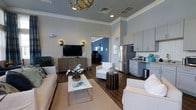 This Great Room and Internet Café is a thoughtfully designed communal space that blends relaxed lounge comfort with functional amenities. The interior showcases a coastal-inspired color palette of soft blues and crisp whites, complemented by rich dark hardwood flooring that grounds the space with warmth and contrast.

The lounge area is anchored by plush white seating arranged around a sleek white coffee table, creating an inviting setting for casual gatherings or quiet relaxation. A wall-mounted flat-screen television sits atop a textured wood media console, flanked by modern wall sconces that add ambient lighting. An elegant gold-toned chandelier overhead serves as a decorative focal point, elevating the overall aesthetic.

Adjacent to the lounge, a well-appointed kitchenette features light grey shaker-style cabinetry, a stainless steel refrigerator, and dark countertops with a classic subway tile backsplash — offering residents convenient access to refreshments. Crown molding details along the ceiling add a refined architectural touch throughout.

A separate workspace area is visible beyond, featuring a bold blue accent wall that visually defines the internet café zone, providing a dedicated environment for productivity. Together, these areas create a versatile, welcoming communal space that balances style with everyday functionality.