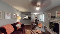 Located at 4720 Reading Rd in Rosenberg, Texas, this single-story home offers a thoughtfully arranged floor plan designed for comfortable everyday living. The living room serves as the heart of the home, featuring rich hardwood-style flooring, a ceiling fan, and a decorative fireplace with a mosaic tile surround and classic white mantle. Large windows dressed with sheer curtains allow natural light to fill the space. The open layout connects the living area to a dedicated dining space, which comfortably accommodates a full dining set and benefits from its own window view. The galley-style kitchen is well-equipped with white cabinetry, granite-style countertops, a double sink, and black appliances including a refrigerator, range, and dishwasher, with a pass-through that opens toward the dining area. The primary bedroom features plush carpeting, a ceiling fan, and natural light, while a second bedroom carries a distinct character with bold nautical-themed accents. Both bathrooms are finished with white cabinetry, granite-style countertops, large vanity mirrors, and shower-tub combinations. Warm gray tones throughout the home create a cohesive and inviting ambiance from room to room.
