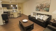 This single-floor residence offers a thoughtfully designed open-concept layout that seamlessly connects the living and kitchen areas. Rich, dark hardwood flooring runs throughout, lending warmth and a cohesive aesthetic to the entire space.

The living area is generously sized, providing ample room for relaxed entertaining and everyday comfort. A large sofa serves as the focal point, complemented by a sturdy wooden coffee table. The neutral-toned walls are accented by decorative artwork, adding character and visual interest to the space.

The kitchen is sleek and functional, featuring modern stainless steel appliances, including a microwave and range. A light-toned breakfast bar with bar-height seating offers a casual dining option and creates a natural transition between the kitchen and living space. Pendant lighting above the counter adds a stylish finishing touch and enhances the overall ambiance.

The open layout and consistent flooring throughout contribute to a sense of spaciousness and flow, making the space feel connected and inviting. The neutral color palette and contemporary finishes create a clean, modern atmosphere that is both practical and visually appealing throughout.