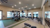 This fitness center is a thoughtfully designed single-floor facility offering a comprehensive workout environment. The space is bathed in natural light streaming through large windows along the perimeter walls, providing views of the surrounding outdoor area and creating an open, energizing atmosphere. Recessed lighting throughout the ceiling supplements the natural light, ensuring the space is well-illuminated at all times. A ceiling fan adds to the comfort of the room by promoting airflow during intense workouts.

The facility is divided into distinct functional zones. A dedicated strength training area features a multi-function cable machine and free weights, including a dumbbell rack, allowing for a wide variety of resistance exercises. A green rubberized flooring section clearly demarcates this zone, providing both safety and durability. The cardio section is well-stocked with treadmills, elliptical machines, and a stationary bike, all positioned to face the windows for an engaging workout experience. A wall-mounted television is also present, adding entertainment options for users. The space maintains a clean, neutral aesthetic with light-toned walls and flooring.