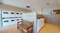This communal laundry facility is designed with both functionality and comfort in mind. The room features a clean, bright aesthetic with recessed lighting set into a white ceiling, ensuring excellent visibility throughout the space. Light beige ceramic floor tiles and tiled wall paneling with a decorative border accent create a neat, low-maintenance environment that is easy to clean and maintain.

The laundry room is well-stocked with multiple stacked washer and dryer units along one wall, maximizing the use of vertical space, while a row of standard top-load washing machines lines an adjacent section, offering users a variety of options to suit their laundry needs. A digital payment or control panel is conveniently positioned near the top-load machines, streamlining the user experience.

A sturdy wooden bench with a classic slatted design is centrally placed in the room, providing a comfortable seating area for those waiting during wash or dry cycles. The overall layout is thoughtfully organized, with clear circulation paths between the equipment. The combination of ample machines, practical finishes, and a welcoming seating area makes this laundry facility a highly functional and user-friendly shared amenity.