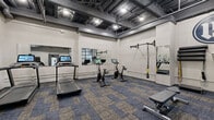 Located at 5820 Patterson Ave in Richmond, VA, this fitness center offers a well-equipped exercise space designed for a comprehensive workout experience. The room features an industrial-style aesthetic, highlighted by an exposed ceiling with visible ductwork, structural beams, and track lighting that bathes the space in bright, functional illumination. The flooring consists of durable patterned carpet tiles in blue and gray tones, providing comfort and traction during exercise.

The cardio section includes a treadmill and multiple Peloton stationary bikes, each equipped with an interactive touchscreen display for guided workouts. A wall-mounted TRX suspension training system offers versatile bodyweight training options, while a dedicated free weights area features a full rack of dumbbells in varying sizes, a BOSU balance trainer, and an adjustable workout bench. Stability balls and medicine ball stacks are also available for functional training.

Large wall-mounted mirrors throughout the space enhance the sense of openness and allow users to monitor their form. A bold accent wall in navy blue displays a prominent logo, adding a branded identity to the space. A water fountain and storage area are conveniently accessible. Floor-to-ceiling black-framed windows along one wall introduce natural light, reinforcing the modern industrial character of this well-appointed fitness facility.