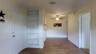 This single-story space features an open layout with warm hardwood-style flooring that runs throughout, lending a cohesive and welcoming feel. The main living area is anchored by a ceiling fan with integrated lighting, providing both comfort and illumination. A standout architectural feature is the built-in floor-to-ceiling shelving unit, offering generous vertical storage and display space. The shelving is finished in crisp white, complementing the neutral, light-toned walls that keep the space feeling bright and airy. A pass-through counter or bar area is visible toward the rear, suggesting a connected kitchen or kitchenette that promotes an open flow between living and cooking spaces. A doorway leads to an additional room, indicating functional zoning within the layout. The clean, minimalist design of the space provides a versatile backdrop suitable for a variety of furnishing styles. HVAC vents are integrated into the ceiling, ensuring climate control throughout. Overall, the space presents a practical and straightforward layout with notable built-in storage and a comfortable, open atmosphere.