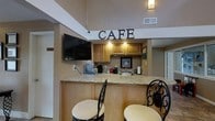 This café space exudes a warm and inviting ambiance, thoughtfully designed for comfort and casual gathering. The focal point of the space is a granite-topped bar counter, accented by decorative wrought-iron bar stools with cushioned seats, offering a welcoming spot for guests to sit and enjoy a beverage or light refreshment. A bold "CAFE" sign adorns the wall above the service area, immediately setting the tone of the space. The service area behind the counter features warm honey-toned wooden cabinetry with under-cabinet lighting, a utility sink, a coffee maker, and a water cooler, providing all the essentials for a fully functional café setup. A wall-mounted flat-screen television is positioned for easy viewing from the counter, adding an entertainment element to the space. The high ceilings with elevated soffits lend a sense of openness and architectural interest, while neutral-toned walls and warm lighting create a relaxed, approachable atmosphere. The overall design blends functionality with comfort, making this café area an ideal spot for informal socializing, refreshments, and day-to-day convenience.