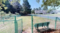 This outdoor space functions as a dedicated dog park or pet exercise area within a residential community. The space is enclosed by a green chain-link fence, providing a secure environment for pets to roam freely. A double-gate entry system is positioned at the front, offering a safe transition point before entering the main enclosure — a common and practical feature in dog parks designed to prevent accidental escapes.

The interior of the fenced area consists of an open, grassy lawn, providing ample room for pets to run and play. The grounds are bordered by mature pine and deciduous trees, which frame the space and offer natural shade on sunny days. A green bench is conveniently placed just outside the entrance gate, giving pet owners a comfortable spot to sit and supervise their animals.

The surrounding environment reflects a well-maintained residential community, with paved roadways, parking areas, and multi-unit residential buildings visible in the background. The overall setting is open and airy, bathed in natural light, with a relaxed, suburban character that complements the community-oriented purpose of the space.
