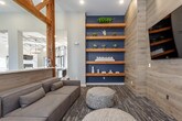 This single-floor amenity space is designed with a sophisticated blend of rustic and modern aesthetics, creating an inviting and functional environment for socializing and recreation. Prominent exposed wooden truss frameworks serve as architectural focal points throughout, adding warmth and a distinctive structural character to the open-concept layout.

The game room area centers around a sleek, full-size billiards table positioned beneath a contemporary cylindrical pendant chandelier. Adjacent to the table is a built-in bar counter with bar-height seating, wood-paneled accents, open shelving, and a wall-mounted television, making it an ideal gathering spot.

The lounge area features a generously sized modular sofa complemented by woven pouf ottomans, creating a relaxed and casual seating arrangement. Deep navy accent walls contrast with lighter tones elsewhere, while floor-to-ceiling curtained windows allow natural light to fill the space. Built-in wooden shelving with decorative objects adds visual interest.

Consistent patterned carpet flooring runs throughout, tying the zones together cohesively. Recessed lighting and pendant fixtures ensure well-distributed illumination, enhancing the overall ambiance of this thoughtfully designed communal space.