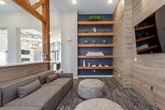 This single-floor amenity space is designed with a sophisticated blend of rustic and modern aesthetics, creating an inviting and functional environment for socializing and recreation. Prominent exposed wooden truss frameworks serve as architectural focal points throughout, adding warmth and a distinctive structural character to the open-concept layout.

The game room area centers around a sleek, full-size billiards table positioned beneath a contemporary cylindrical pendant chandelier. Adjacent to the table is a built-in bar counter with bar-height seating, wood-paneled accents, open shelving, and a wall-mounted television, making it an ideal gathering spot.

The lounge area features a generously sized modular sofa complemented by woven pouf ottomans, creating a relaxed and casual seating arrangement. Deep navy accent walls contrast with lighter tones elsewhere, while floor-to-ceiling curtained windows allow natural light to fill the space. Built-in wooden shelving with decorative objects adds visual interest.

Consistent patterned carpet flooring runs throughout, tying the zones together cohesively. Recessed lighting and pendant fixtures ensure well-distributed illumination, enhancing the overall ambiance of this thoughtfully designed communal space.