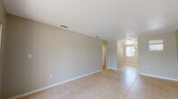 This single-story residence offers a clean, functional layout with consistent light beige ceramic tile flooring running throughout every room, creating a cohesive and easy-to-maintain living environment. Neutral warm-gray walls and white baseboards complement the textured ceilings, giving the interior a simple, understated character.

The open living area is generously proportioned, with views through to a bright dining space illuminated by a pendant light fixture and a window that allows ample natural light to filter in. A sliding glass door in the adjoining room opens to a balcony overlooking lush tropical landscaping and what appears to be a pool area, offering pleasant outdoor views framed by palm trees.

The kitchen is equipped with white cabinetry, marble-patterned countertops, a white electric range with an overhead exhaust hood, and a white refrigerator. A classic subway tile backsplash adds a clean, timeless detail to the cooking area.

The bedroom features a large window with views of tropical greenery, filling the space with natural light. Multiple closet doors provide ample storage within the room. The bathroom includes a full-size soaking tub with an overhead shower, built-in shelving, and tile flooring, finished in a clean, all-white palette. Central HVAC vents are present throughout, ensuring year-round comfort.