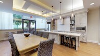 This single-floor communal space seamlessly blends a fully equipped kitchen with a generous dining area, creating an inviting environment designed for both functionality and elegance. The kitchen is appointed with crisp white cabinetry, open display shelving stocked with cookbooks and dishware, a stainless steel French door refrigerator, a built-in microwave, and a cooktop with a sleek wall-mounted range hood. A long, white-topped island with turned legs provides ample prep space and casual seating with dark wood bar stools, all illuminated by a row of glass pendant lights that add a refined touch overhead. The dining area accommodates a large, light wood dining table surrounded by patterned upholstered chairs in blue and white, perfect for group meals or meetings. A wall-mounted flat-screen TV is flanked by a curated gallery of framed artwork, adding a polished, residential feel. The space is finished with warm light hardwood flooring throughout, a coffered ceiling with recessed and cove lighting, and large glass doors that open to an outdoor patio, filling the interior with natural light and greenery views. The overall ambiance is sophisticated yet welcoming, offering a versatile setting for dining, socializing, and collaborative gatherings.