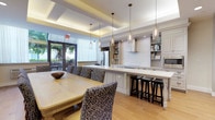 This single-floor communal space seamlessly blends a fully equipped kitchen with a generous dining area, creating an inviting environment designed for both functionality and elegance. The kitchen is appointed with crisp white cabinetry, open display shelving stocked with cookbooks and dishware, a stainless steel French door refrigerator, a built-in microwave, and a cooktop with a sleek wall-mounted range hood. A long, white-topped island with turned legs provides ample prep space and casual seating with dark wood bar stools, all illuminated by a row of glass pendant lights that add a refined touch overhead. The dining area accommodates a large, light wood dining table surrounded by patterned upholstered chairs in blue and white, perfect for group meals or meetings. A wall-mounted flat-screen TV is flanked by a curated gallery of framed artwork, adding a polished, residential feel. The space is finished with warm light hardwood flooring throughout, a coffered ceiling with recessed and cove lighting, and large glass doors that open to an outdoor patio, filling the interior with natural light and greenery views. The overall ambiance is sophisticated yet welcoming, offering a versatile setting for dining, socializing, and collaborative gatherings.