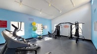 This fitness center offers a comprehensive workout environment designed to support a wide range of exercise routines. The space features a high, vaulted ceiling that enhances the open and airy atmosphere, complemented by track lighting that ensures the entire area is well-lit throughout the day. Large windows allow natural light to filter in, creating an inviting and energizing environment for workouts.

The floor is finished with durable rubber flooring, providing both safety and comfort during exercise. The space is equipped with a variety of cardio machines, including a treadmill and an elliptical, positioned to take advantage of the natural light. A multi-station cable machine anchors the strength training area, offering users a versatile range of resistance exercises. An adjustable workout bench adds further flexibility for strength routines.

A vibrant blue accent wall adds a dynamic visual element to the space and serves as the backdrop for a wall-mounted storage rack holding an assortment of stability balls and resistance equipment. Motivational wall decals reinforce the energetic atmosphere of the room. The overall design balances functionality with an uplifting aesthetic, making this fitness center a practical and welcoming space for daily exercise.