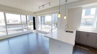 This modern urban residence offers a thoughtfully designed open-concept layout that seamlessly blends the living and kitchen areas into a cohesive, light-filled space. The expansive floor-to-ceiling windows are a defining architectural feature, flooding the interior with abundant natural light while framing sweeping views of the surrounding cityscape, including palm trees and neighboring buildings that speak to a vibrant urban setting.

The kitchen is a standout element, featuring a generous waterfall-edge island with a pristine white stone countertop that serves as both a functional workspace and a casual gathering spot. A stainless steel undermount sink with a contemporary arc faucet adds a refined touch, while stylish pendant lights hang above, providing warm task lighting. Dark cabinetry below the island provides a striking contrast to the bright countertops and white walls.

The living area is anchored by rich, dark hardwood-style flooring that adds warmth and depth to the otherwise crisp, neutral palette. Overhead, a sleek track lighting system provides flexible, adjustable illumination. The overall ambiance is clean, contemporary, and airy — a sophisticated urban retreat that balances minimalist aesthetics with functional, modern living.