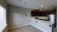 This single-story space showcases a seamlessly integrated kitchen and dining area designed with a clean, modern aesthetic. The kitchen is equipped with rich, dark espresso-toned cabinetry that contrasts sharply against the bright white countertops, creating a striking visual balance. A peninsula with an overhang provides casual seating and doubles as a functional workspace, while a stainless steel sink and gas range add practicality to the layout. A built-in microwave is neatly integrated into the upper cabinetry, maintaining the streamlined appearance.

Recessed lighting illuminates the kitchen area efficiently, while a brushed nickel pendant light hangs above the adjacent dining space, serving as a focal point and providing warm, directed light. The flooring throughout features light wood-look tile, offering both durability and a warm, neutral foundation that complements the overall color palette. Neutral gray walls contribute to the airy, contemporary feel of the space. White baseboards provide a clean finishing detail throughout. The open-concept layout between the kitchen and dining area promotes an easy flow and a sense of spaciousness, making the space both functional and inviting.
