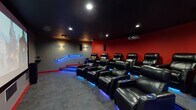 Located at 150 Southfield Ave in Stamford, Connecticut, this dedicated private theatre delivers an immersive cinematic experience within a thoughtfully designed single-floor space. The room is anchored by a large projection screen that dominates the front wall, flanked by floor-standing speakers and complemented by a ceiling-mounted projector, together delivering a powerful audiovisual setup well-suited for film screenings and private events.

Seating is arranged in tiered rows, allowing unobstructed sightlines from every position. Each seat is a plush black leather power recliner equipped with built-in cup holders and integrated blue LED lighting at the base, lending the space a dramatic, cinematic atmosphere. The tiered platform design, accented by blue LED-lit handrails along the steps, reinforces the theatre's professional layout.

The interior design draws on bold contrasts: deep red accent walls curve along the seating area, while grey walls frame the screen and entrance zones. Acoustic panels are strategically mounted throughout to optimize sound quality and minimize reverberation. Recessed ceiling lights and wall sconces provide subtle, controlled illumination that enhances the viewing ambiance without detracting from the screen. Dark carpeting and a dark ceiling complete the immersive, light-controlled environment, making this theatre a premier destination for an elevated screening experience.