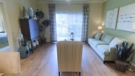 This single-floor living space offers a well-appointed interior centered around a bright and airy main living area. The room features warm hardwood-style flooring that runs throughout, lending a clean and cohesive feel to the space. A large sliding glass door dominates the far wall, fitted with vertical blinds and framed by patterned draping curtains that add a decorative touch while allowing for flexible light control. Natural light flows generously into the room, creating an open and welcoming ambiance. The living area is comfortably furnished with a light-colored sofa accented by striped throw pillows, and a striped accent chair positioned centrally, offering a functional seating arrangement. A tall decorative plant in the corner adds a natural element and visual height to the space. To one side, a dark cabinet with diamond-patterned detailing provides storage and display space, with decorative items arranged on top. A softly lit lamp on the opposite side contributes to the warm, layered lighting. The overall aesthetic is casual yet thoughtfully styled, making the space feel comfortable, livable, and inviting.